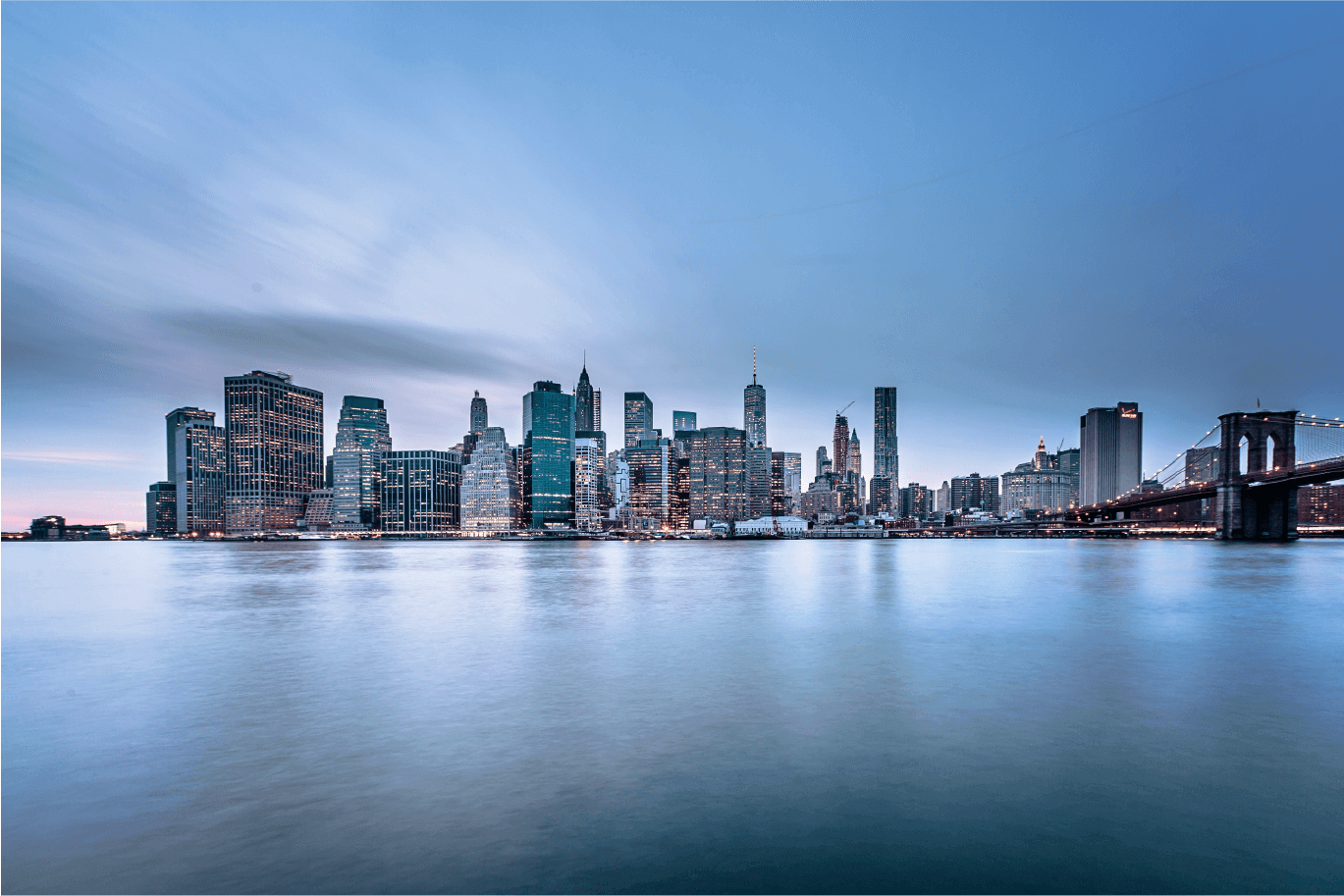 Wide panoramic shot of the Lower Manhattan skyline and the Brooklyn Bridge at dusk.