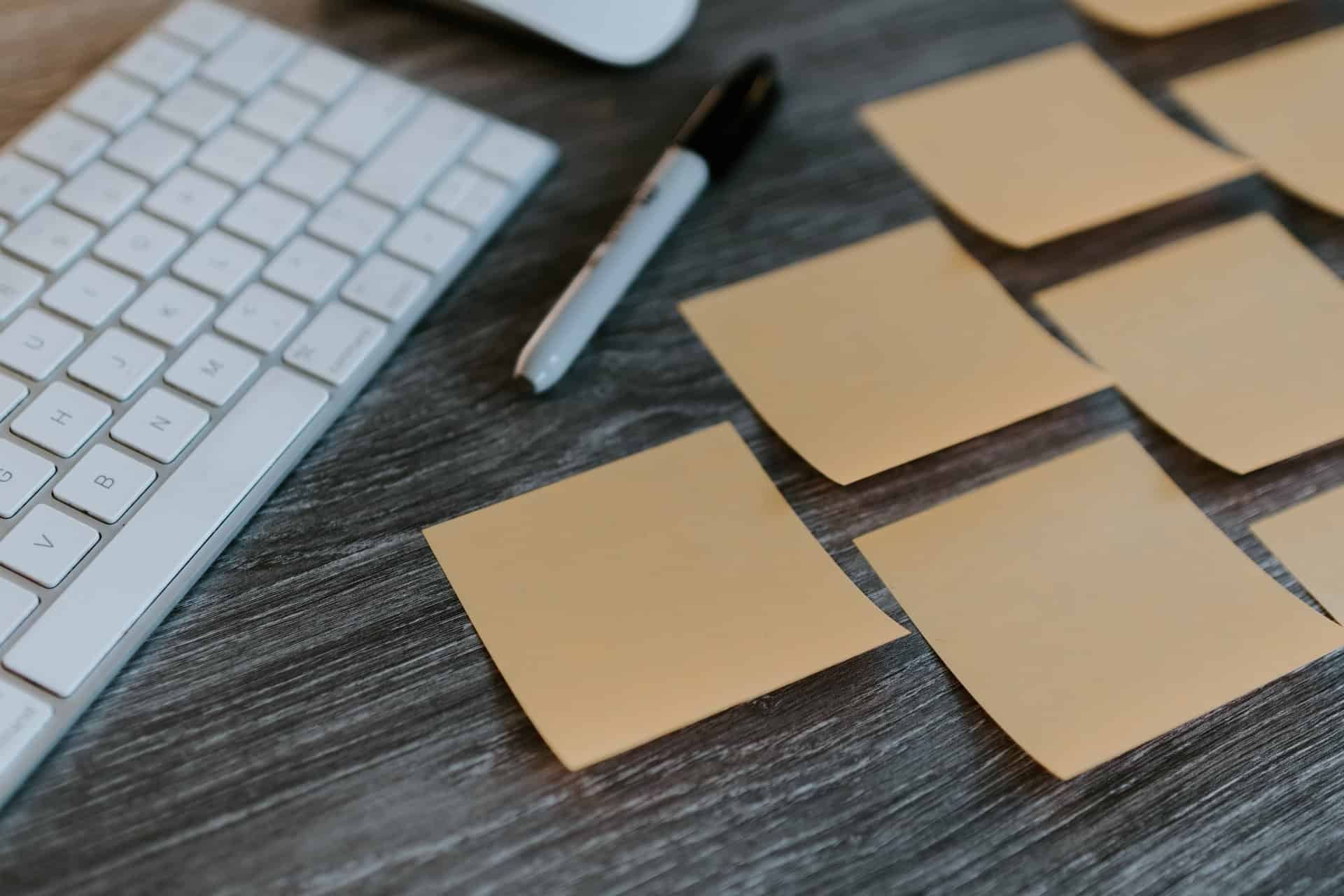Close-up of beige sticky notes scattered on a dark wooden desk next to a white keyboard and black marker.
