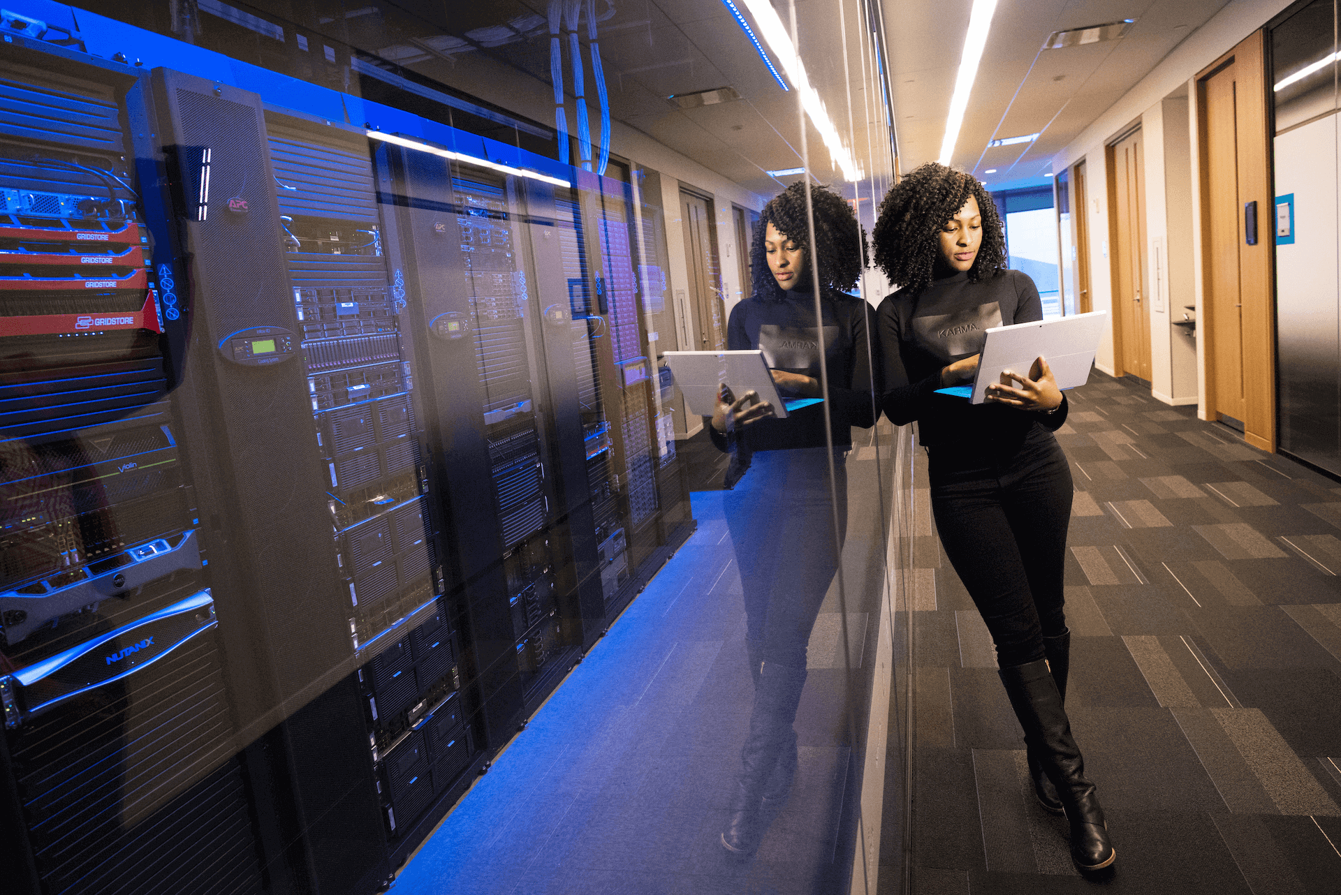 Woman standing next to a glass wall, holding a tablet, with server racks visible behind the glass.