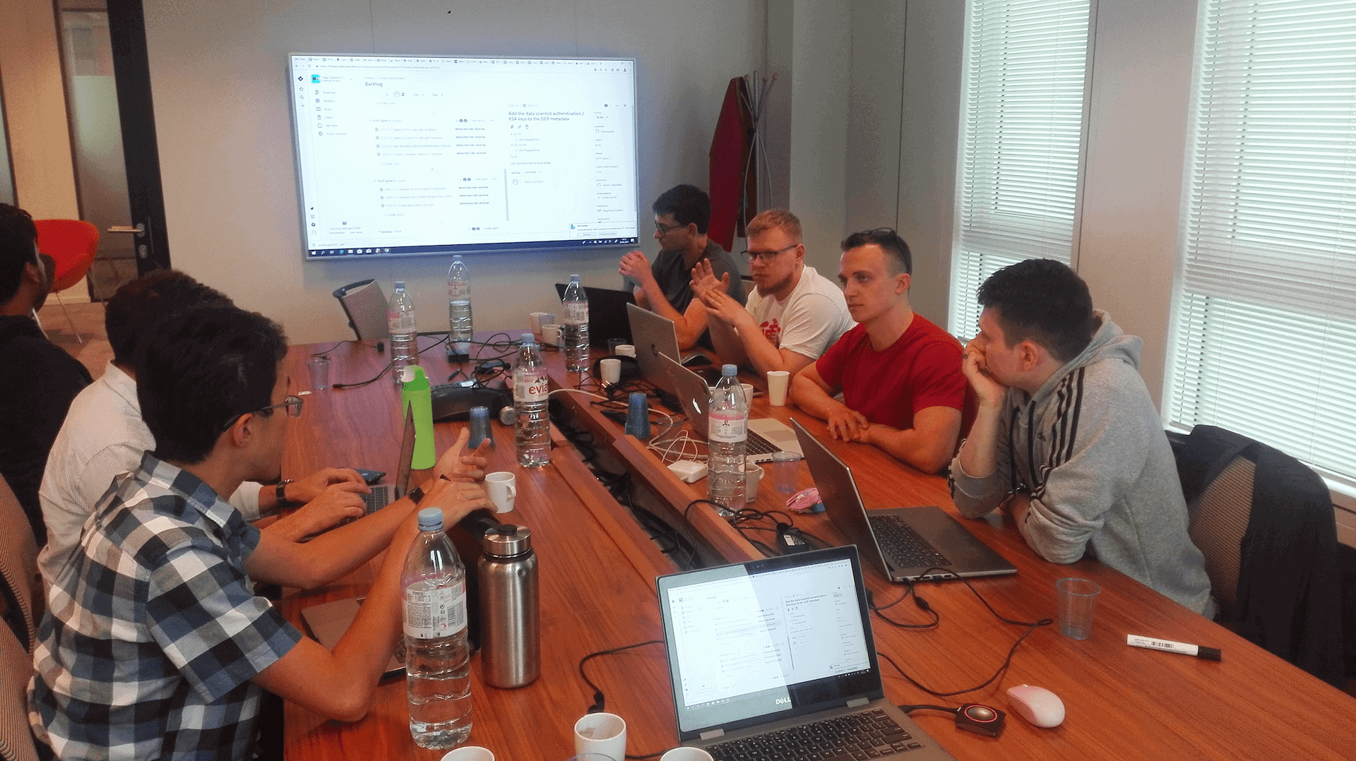 Group of men in a meeting seated around a long table with laptops and a large screen displaying data.