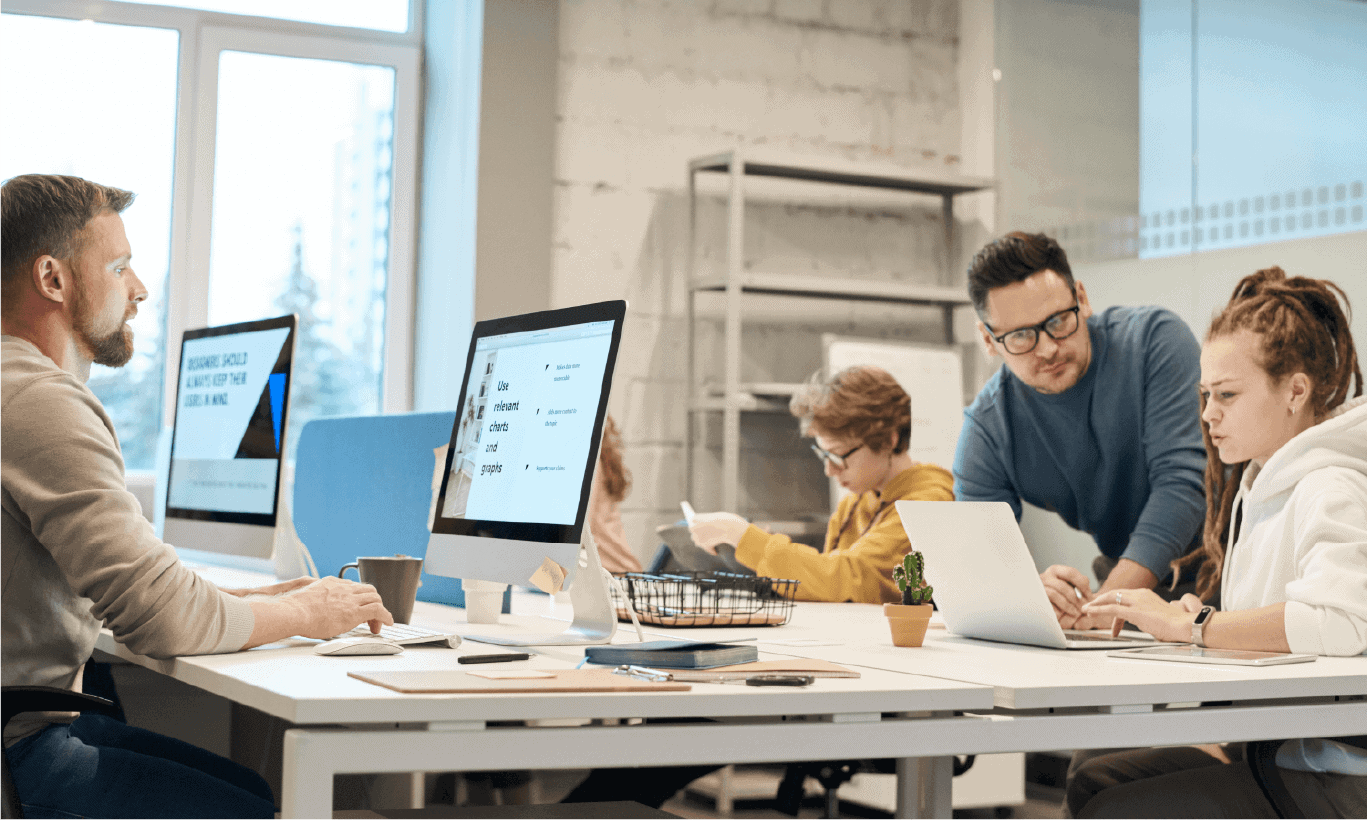Four people working collaboratively at a white desk in a modern, bright office.