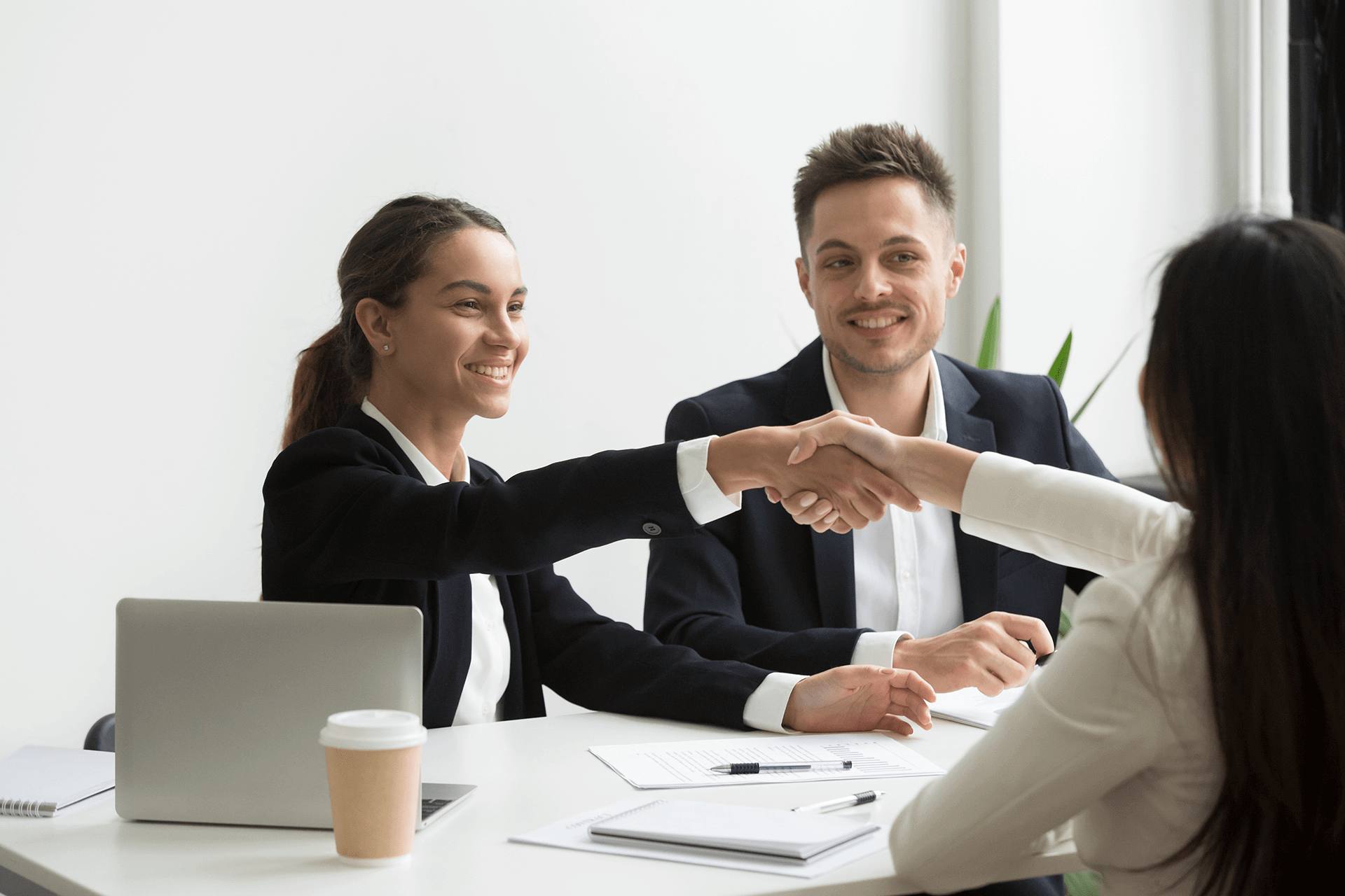 Two professionals shaking hands across a white table during a business meeting.