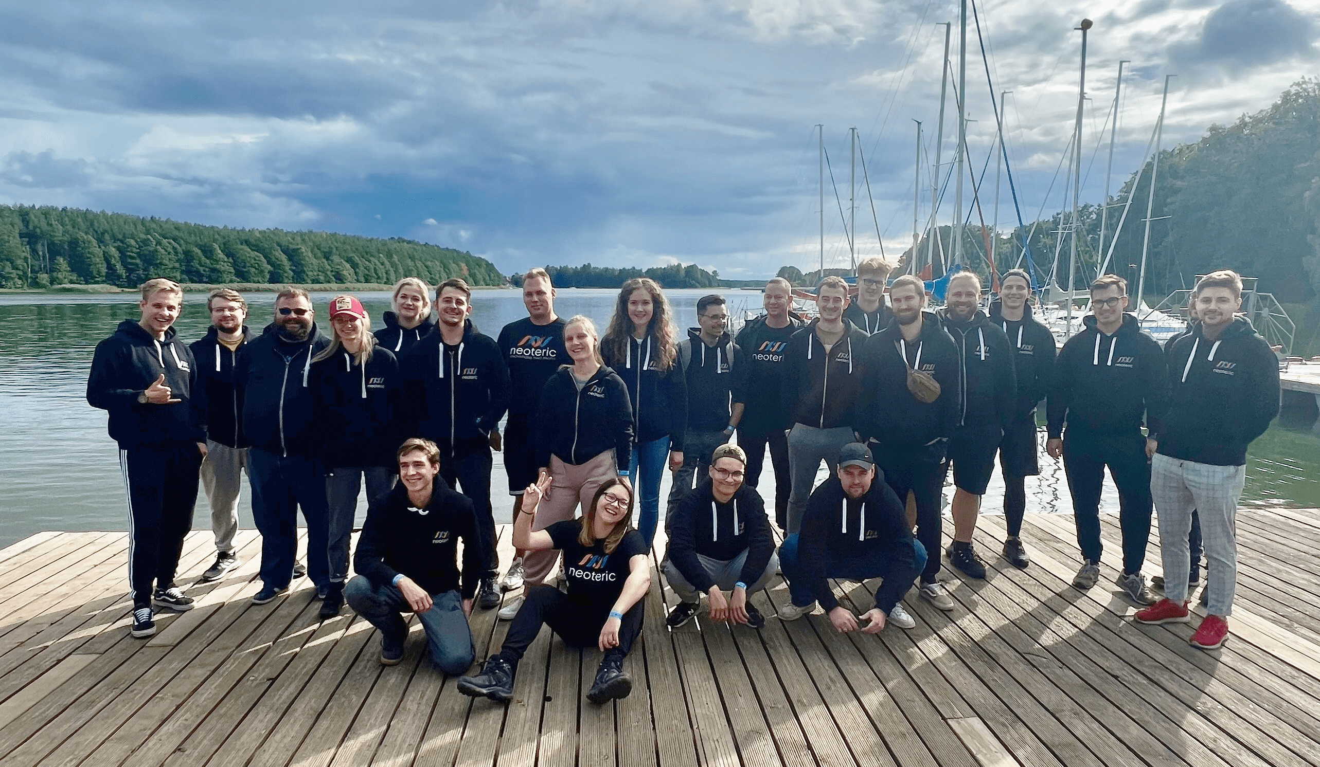 Group of people in matching hoodies posing on a lakeside dock with sailboats and forested hills in the background.